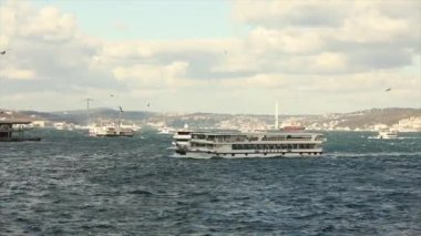 Boğaz köprüsünün arka planında tekneler, rüzgarlı hava. Boğaziçi Köprüsü, ön planda turistler ve martılarla dolu eğlence tekneleri..