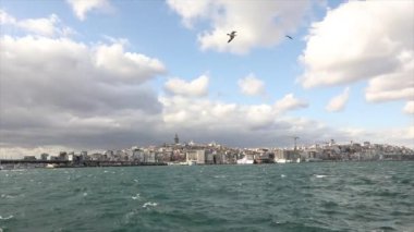 Galata Kulesi, ön planda turistler ve martılarla dolu eğlence tekneleri. Galata Kulesi 'nin arka planında tekneler, rüzgarlı hava