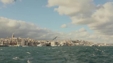 Galata Kulesi 'nin yavaş çekim görüntüleri, ön planda turistler ve martılarla dolu eğlence tekneleri. Galata Kulesi 'nin arka planında tekneler, rüzgarlı hava