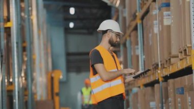 Modern bir depoda bir adam. Depo müdürü depoyu kontrol ediyor. Depoda kask takan bir işçi.