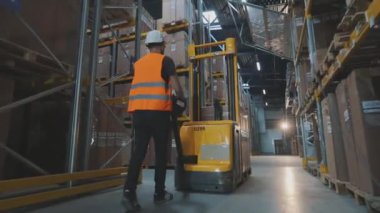 Bir işçi elektrikli forkliftle depoya doğru yürür. Bir işçi depodan kargo taşıyor.