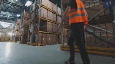 Depoda forklift kullanan bir işçi. İşçi depoya doğru yürür. Depoda çalışan bir işçi elektrikli forklift taşır.