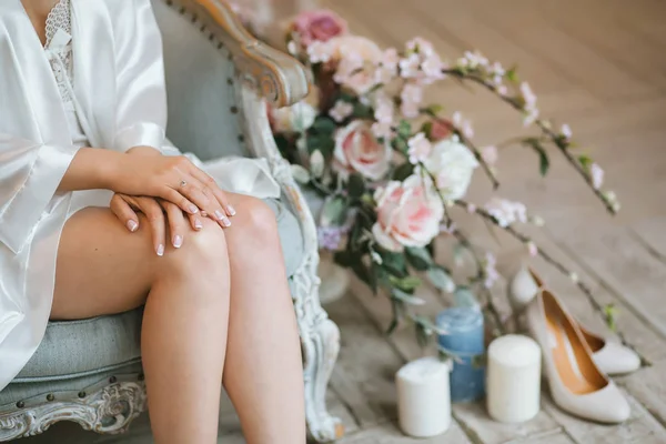 Gelin Dantel ile ipek sabahlık giymiş bir vintage mavi sandalye sabahı oturur, kucağında güzel Fransız manikür katlanmış eller. Yakınlarda bir çiçek buketi, mumlar ve bej Ayakkabı bulunmaktadır