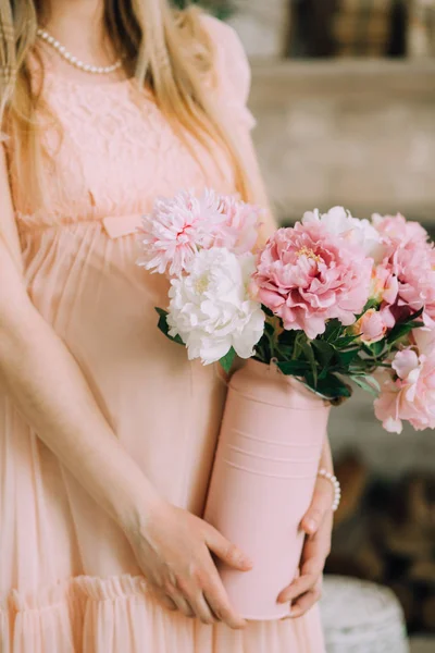 Hamile bir kadın narin pembe elbise ve bir inci kolye peonies vazo ellerini tutar.