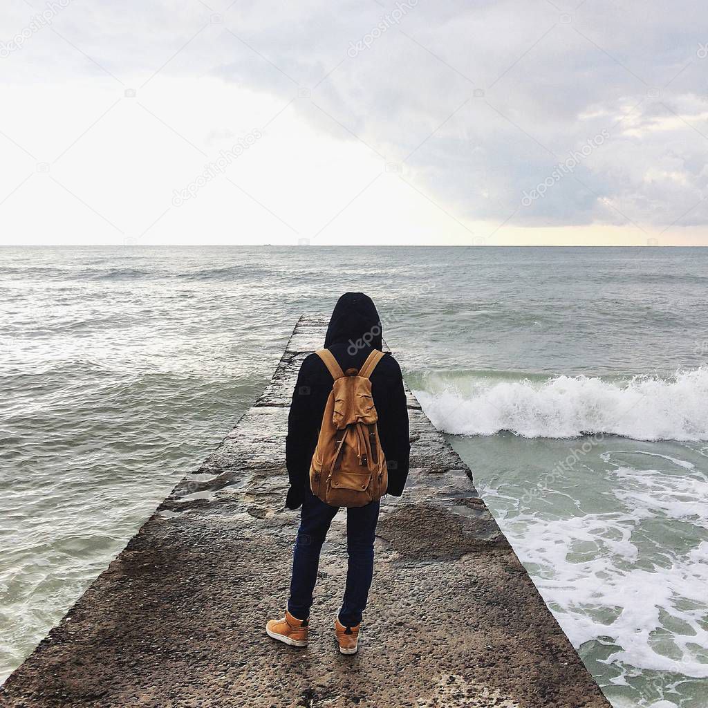 A man with a brown backpack stands with his back on the pier and looks at the storm in the sea. Square