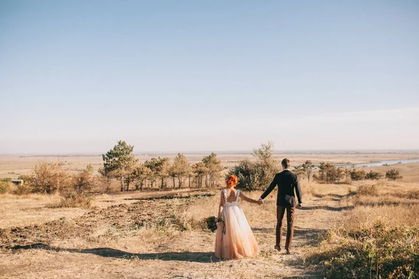 A noiva e o noivo em um vestido de noiva laranja brilhante andam de mãos dadas em uma colina ensolarada com uma bela vista do rio e da floresta no outono Imagens De Bancos De Imagens Sem Royalties
