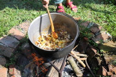 Romanya'da kırsal bir evin arka bahçesinde açık günlük ateşte nerede bir gulaş güveç bir dökme demir kazanda karıştırarak kadın kaynıyor