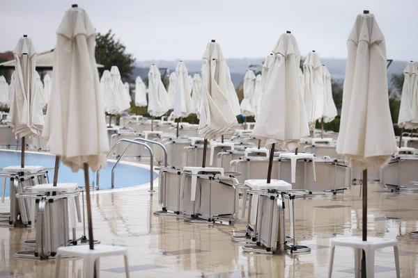 Closed sun umbrellas and lawn chairs around a hotel pool in off season, with no people in a cold cloudy day, in late autumn on the Bulgarian seaside