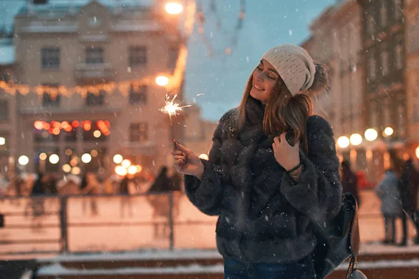 Bella giovane donna in pelliccia in possesso di una scintilla gode di umore natalizio invernale nella vecchia città europea innevata con pista di pattinaggio all'aperto su sfondo — Foto Stock