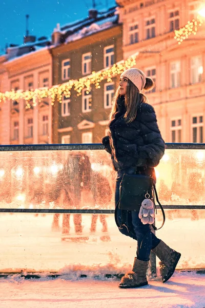 Beautiful young woman in fur coat stands by a skating rink border on old snowy European city background — Stock Photo, Image