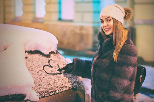 Young beautiful happy woman draws a heart shape symbol on the frozen car windscreen covered with snow — Stock Photo, Image