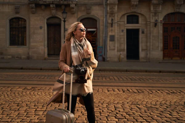 Joven hermosa turista rubia con una bolsa de viaje con ruedas y cámara de cine vintage de pie en la plaza Rynok en Lviv, Ucrania por la mañana — Foto de Stock