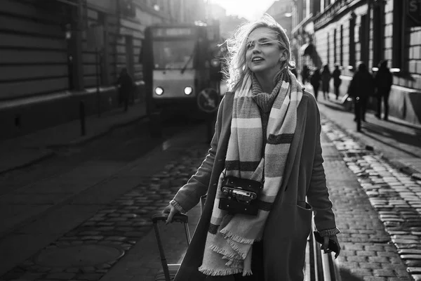 Jovem mulher loira bonita turista com um saco de viagem com rodas e câmera de filme vintage chega à nova cidade para explorá-lo em preto e branco — Fotografia de Stock