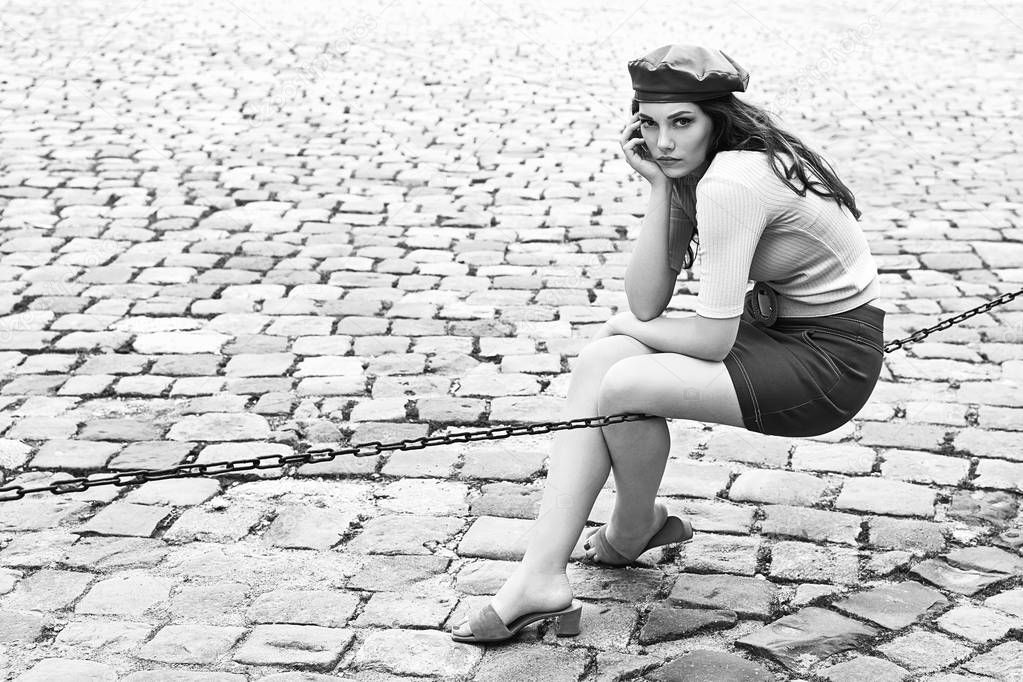 Young beautiful brunette emotional girl dressed in retro vintage style in the old european city sits of a chain