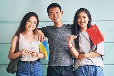Group of three happy international chinese asian students smiling and holding flags of China and Ukraine in university clipart