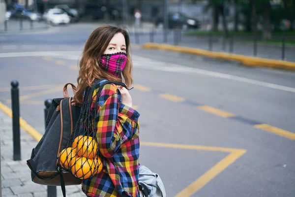 Kockás, kézzel készített arcmaszkot viselő fiatal nő, színes ingben, narancsokkal teli hálós táskával és az utcán álló hátizsákkal.. — Stock Fotó