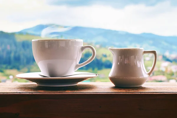 Piani dopo la quarantena. Caffè con latte in tazza di ceramica con piccola brocca di latte sul vecchio davanzale di legno con sfondo montagne estive . — Foto Stock