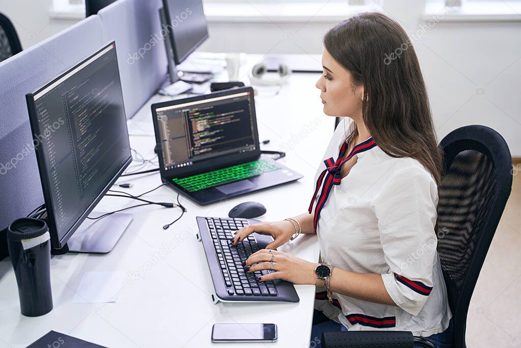 Beautiful female junior software developer working on computer in IT office, sitting at desk and coding, working on a project in software development company or technology startup. High quality image.