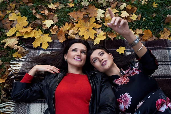 Dos hermosas hermanas adultas están acostadas en una alfombra a cuadros en el parque de otoño con hojas amarillas y naranjas caídas mirando las nubes en el cielo soñando y hablando — Foto de Stock