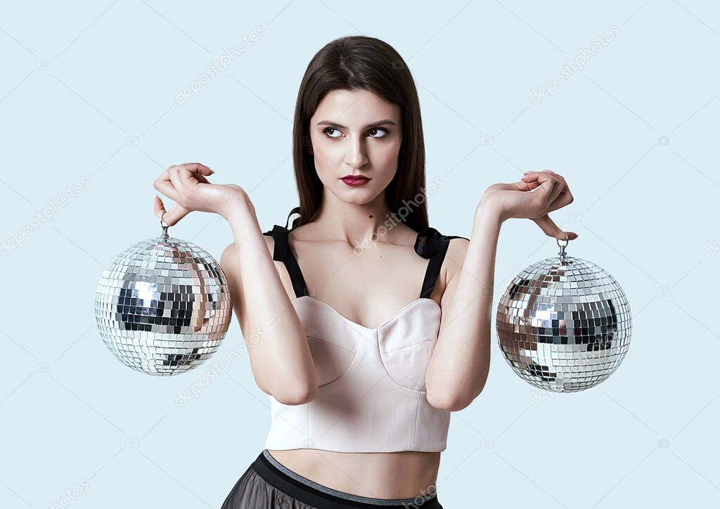 Young beautiful brunette girl posing in studio. Woman balancing holding two disco balls reflecting light.