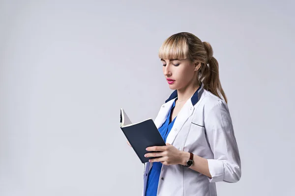 Médica jovem confiante em pé com caderno ou livro médico em suas mãos. Cheques registros, receitas ou prescrições. Estagiário em roupas médicas brancas posando em fundo cinza claro. — Fotografia de Stock