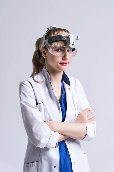 Confident ENT doctor wearing surgical headlight head light and protective glasses. Portrait of female otolaryngologist or head and neck surgeon standing arms crossed on light grey background.