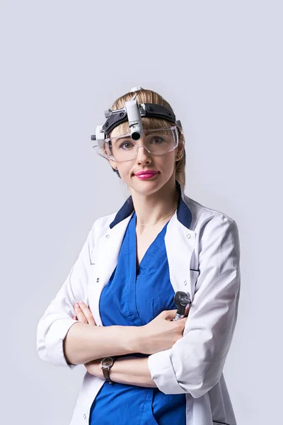 Confident ENT doctor wearing surgical headlight head light and protective glasses. Portrait of female otolaryngologist or head and neck surgeon standing arms crossed on light grey background.