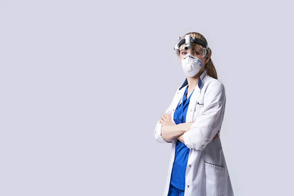 Confident ENT doctor wearing surgical headlight head lantern, protective face mask and glasses. Portrait of female otolaryngologist standing arms crossed on light grey background.