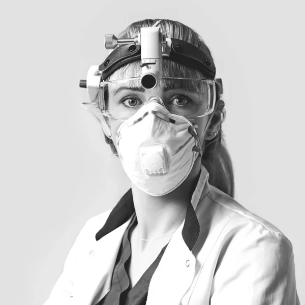 Confident ENT doctor wearing surgical headlight head light and protective glasses. Portrait of female otolaryngologist or head and neck surgeon on light grey background.