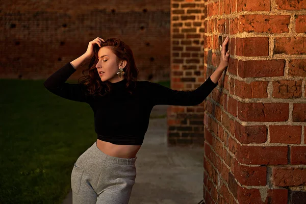 Retrato de uma linda menina ruiva sensual posando ao pôr-do-sol usando luz cinza claro perna larga calças de cintura alta jersey e t-shirt manga longa preta lançando uma sombra na parede de tijolo vermelho — Fotografia de Stock