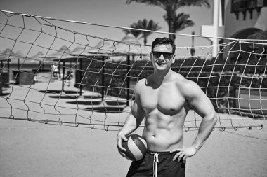 Yüzme şortlu yakışıklı, güçlü atletik genç adam voleybol topunu bir deniz tatil köyünde voleybol sahasında tutuyor. Spor mankeni ya da tatildeki sporcu. Siyah ve beyaz.