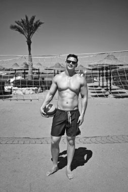 Yüzme şortlu yakışıklı, güçlü atletik genç adam voleybol topunu bir deniz tatil köyünde voleybol sahasında tutuyor. Spor mankeni ya da tatildeki sporcu. Siyah ve beyaz.