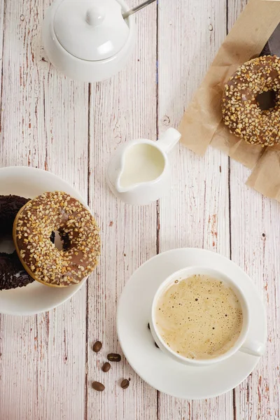 Morning breakfast with chocolate donuts and  cup of coffee with cream on  light wooden background.Delicious donuts sprinkled with crushed nuts.  top view
