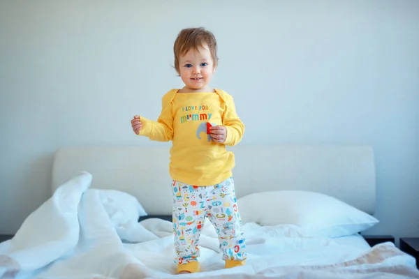 Grappige babymeisje permanent in bed van de ouders. Baby meisje in gele pyjama 's — Stockfoto