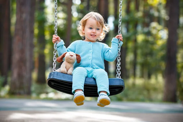 Adorable toddler girl with soft toy dog having fun on a swing on summer day — Stock Photo, Image