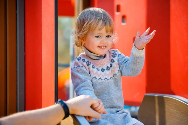 Sweet blond little baby girl in beautiful knitted dress plays outdoors at playground. Father holding a hand of his daughter — Stock Photo, Image