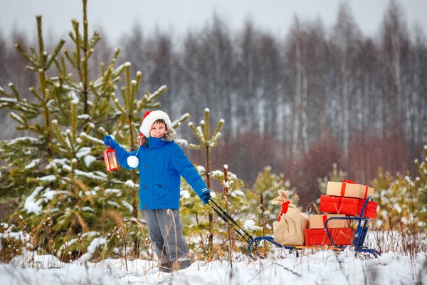 Ładny mały chłopiec w Santa hat prowadzi drewniane sanki z darów w śnieżnym lesie — Zdjęcie stockowe