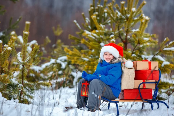 Menino bonito em chapéu de Santa carrega um trenó de madeira com presentes na floresta nevada — Fotografia de Stock