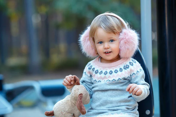 Retrato de doce loira pequena menina em belo vestido quente e inverno faux pêlo orelha muffs no parque infantil — Fotografia de Stock