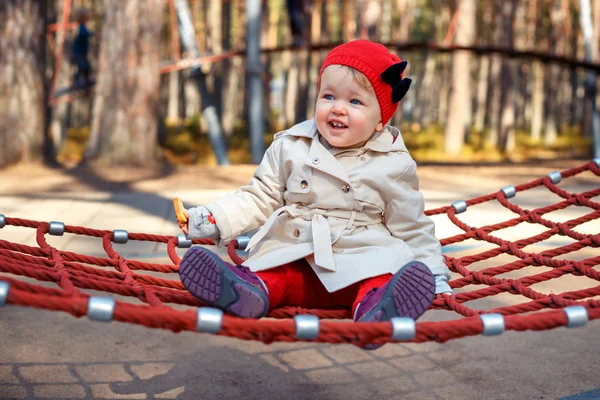 Sweet blond little toddler girl in beautiful beige trench coat and red cap having fun on a swing at playground — Stock Photo, Image