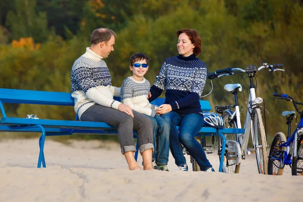 Ung familj på tre med resten på bänken under deras cykel ridning på stranden — Stockfoto