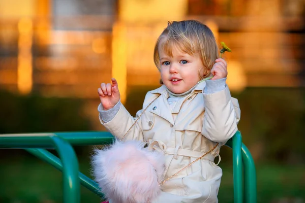 Retrato de doce loira pequena menina em casaco de trincheira e saco de pele falsa no parque infantil — Fotografia de Stock