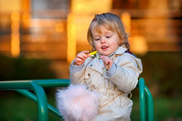 Retrato de doce loira pequena menina em casaco de trincheira e saco de pele falsa no parque infantil — Fotografia de Stock