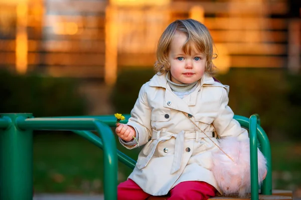 Retrato de doce loira pequena menina em casaco de trincheira e saco de pele falsa no parque infantil — Fotografia de Stock