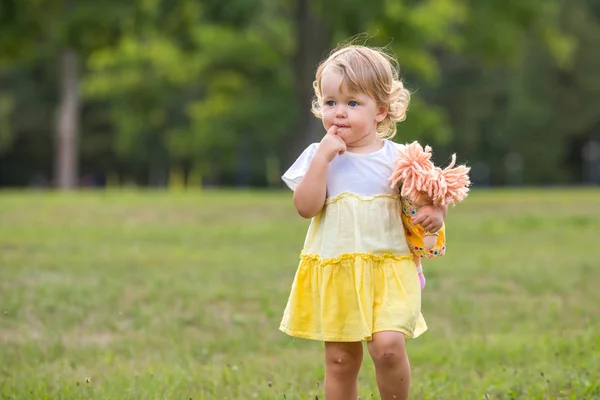 Dolce bionda bambina con dito in bocca e bambolina di pezza imbottita a maglia che si diverte a camminare all'aperto — Foto Stock