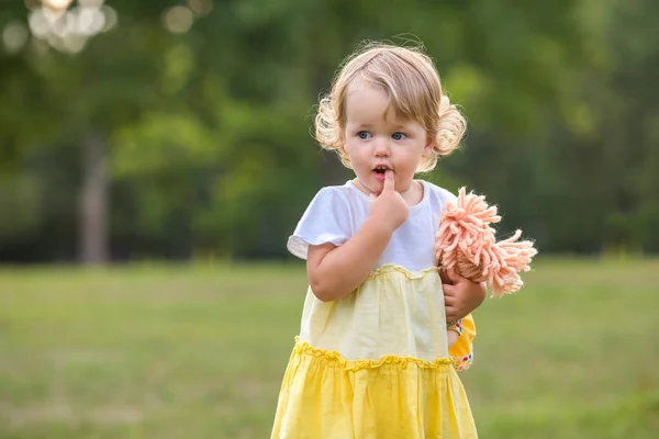 Sweet blond toddler girl with finger in mouth and knitted stuffed rag doll having fun walking outdoors — Stock Photo, Image