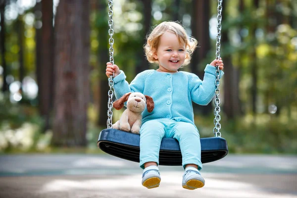 Adorabile bambina con cane giocattolo morbido che si diverte su un'altalena nella giornata estiva — Foto Stock