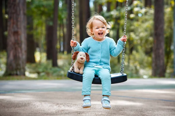 Adorable toddler girl with soft toy dog having fun on a swing on summer day — Stock Photo, Image