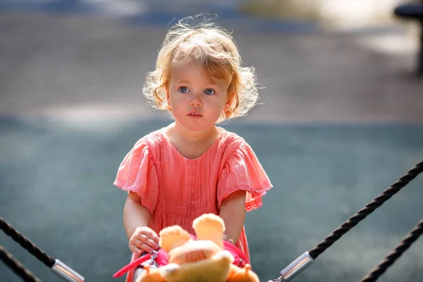 Doce loira pequena menina em belo vestido de damasco joga passeios ao ar livre no parque infantil — Fotografia de Stock