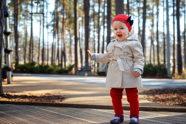 Tatlı sarışın küçük yürümeye başlayan çocuk kız güzel bej trençkot ve kırmızı bere açık havada oyun alanı çalış — Stok fotoğraf
